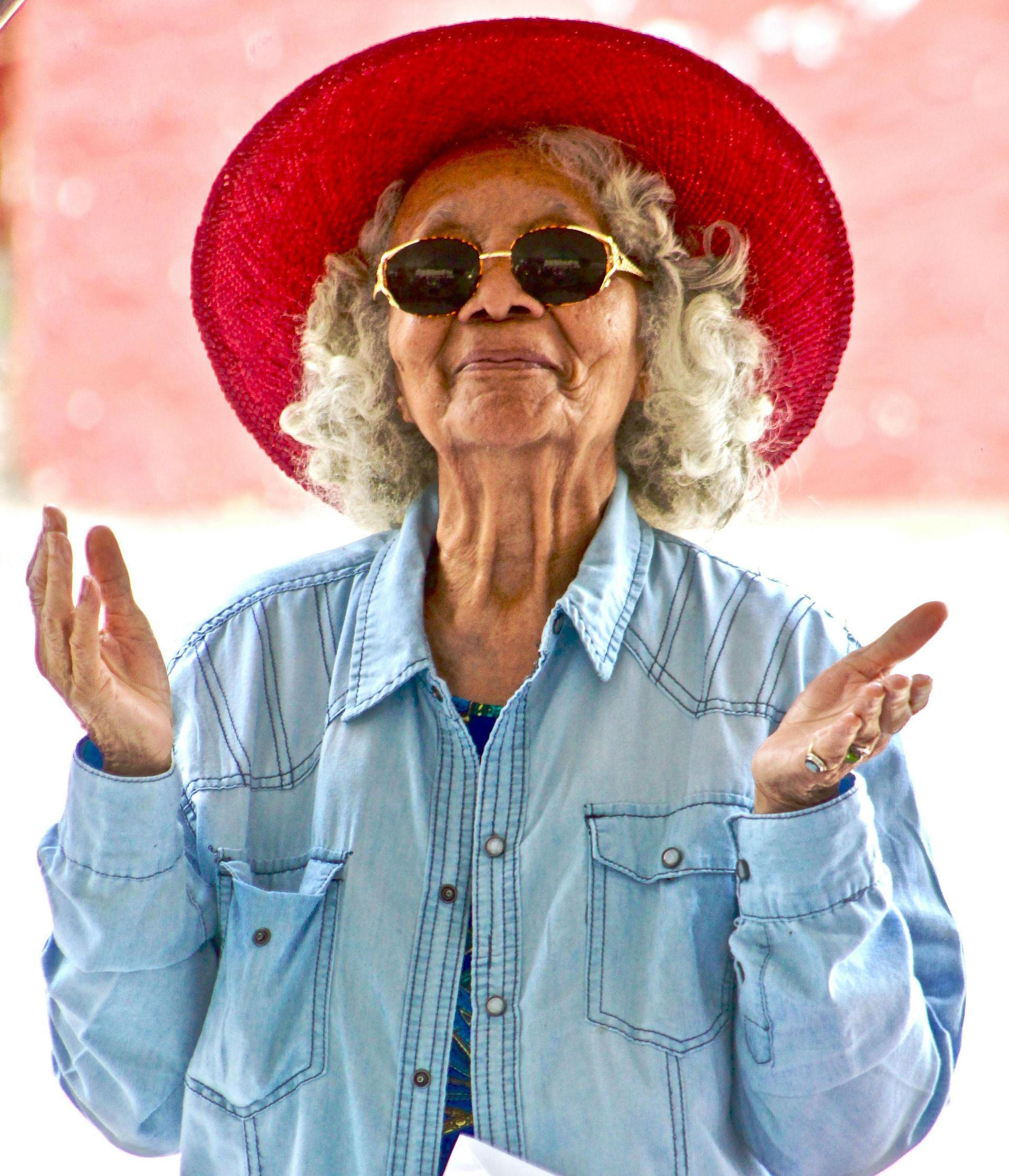Elderly woman in denim shirt, red hat, and sunglasses with open hands, smiling.